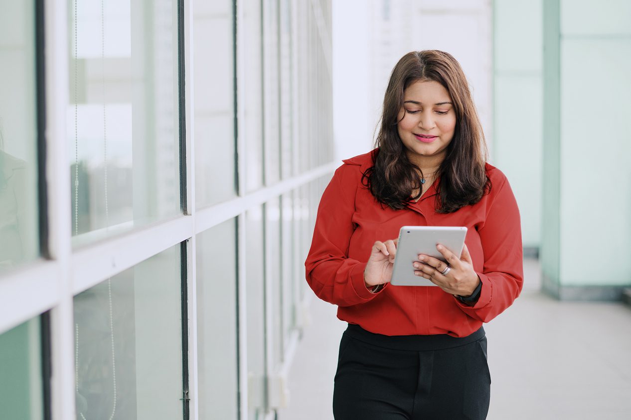 Person in a red blouse standing in a bright office hallway, looking at a tablet screen while interacting with it, near a row of glass windows.