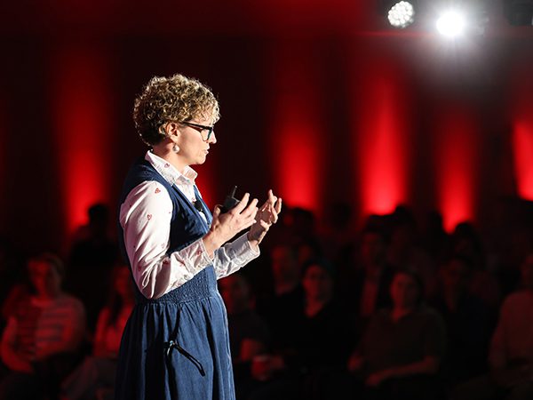 Vanguard_Social_WebContent_UnlimITed_Image03_600x450 Crew presenter on stage at the unlimITed conference, with dramatic lighting featuring spotlights and red perimeter lighting.