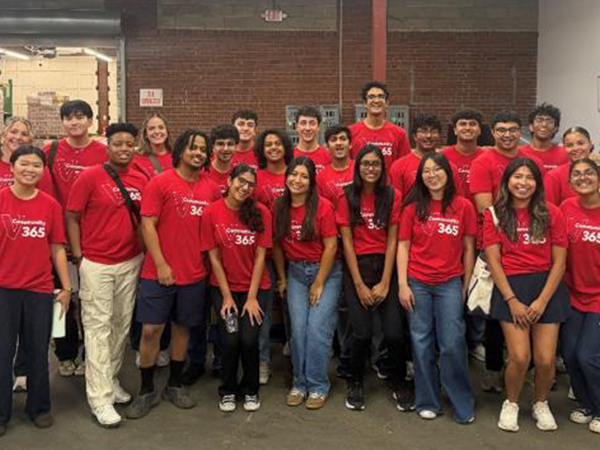 : A large group of Vanguard interns wearing red Community 365 volunteer T-shirts poses for a group photo inside a warehouse during a community service event. Pallets of canned goods and other donated items are visible in the background, along with warehouse shelving and industrial lighting.