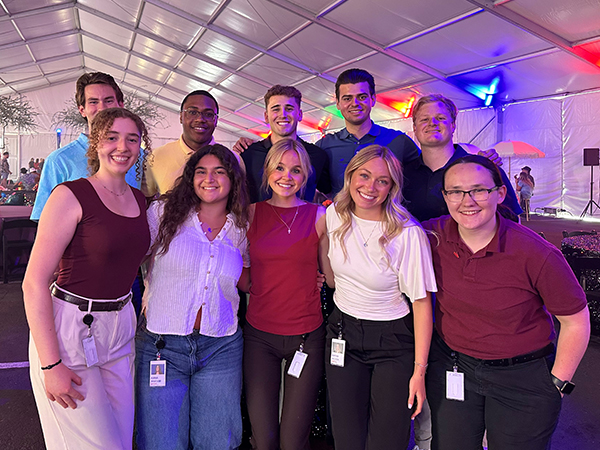 A group of Vanguard interns poses together at an indoor summer event held inside a large tent. The interns wear business casual attire and stand in front of tables and event decorations illuminated by colorful red, blue, and purple lighting. The group gathers for a photo during a social or networking activity as part of the Vanguard internship program