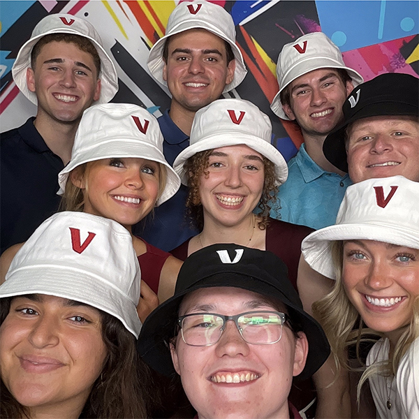 : A group of Vanguard interns poses together for a photo wearing white and black bucket hats featuring a red Vanguard "V" logo. The group stands in front of a colorful, abstract backdrop with bright geometric shapes and lines. The photo captures the interns gathered closely together during a summer internship event or celebration