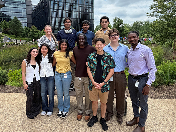 A group of Vanguard interns stands together outdoors in a landscaped courtyard on a Vanguard campus. The group poses in front of modern office buildings, trees, and garden greenery under a cloudy sky. The interns wear a mix of business casual and casual attire and are gathered for a group photo during their summer internship experience