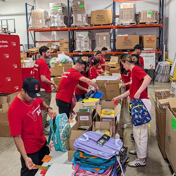 Vanguard interns wearing red Community 365 T-shirts volunteer by sorting and organizing school supplies and backpacks at long tables inside a warehouse space. Shelves filled with boxes and supplies line the background as volunteers work together preparing materials for local students and families.