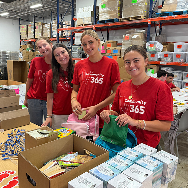 Four Vanguard interns wearing red Community 365 volunteer T-shirts stand together in a warehouse space while assembling backpacks and school supply kits. Tables in front of the group contain backpacks, notebooks, pens, and other supplies, while shelves stocked with boxes and materials are visible in the background.