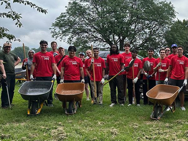 A group of Vanguard interns wearing red Community 365 volunteer T-shirts stands together outdoors holding shovels and standing beside wheelbarrows during a community service event. The volunteers are gathered on a grassy field near a green canopy tent under an overcast sky, preparing to participate in a landscaping or environmental stewardship project
