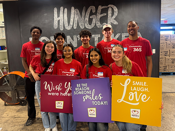 : A group of Vanguard interns wearing red Community 365 volunteer T-shirts pose together. Several interns hold colorful signs that read “Wish you were here!,” “Be the reason someone smiles today!,” and “Smile. Laugh. Love.” A large wall behind the group displays the words “Hunger Can't Wait."