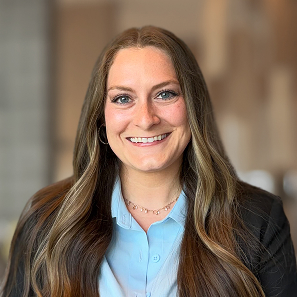 Professional headshot of a person with long brown hair wearing a light blue collared shirt, a dark blazer, hoop earrings, and a delicate necklace. The person is centered in the frame against a softly blurred indoor background
