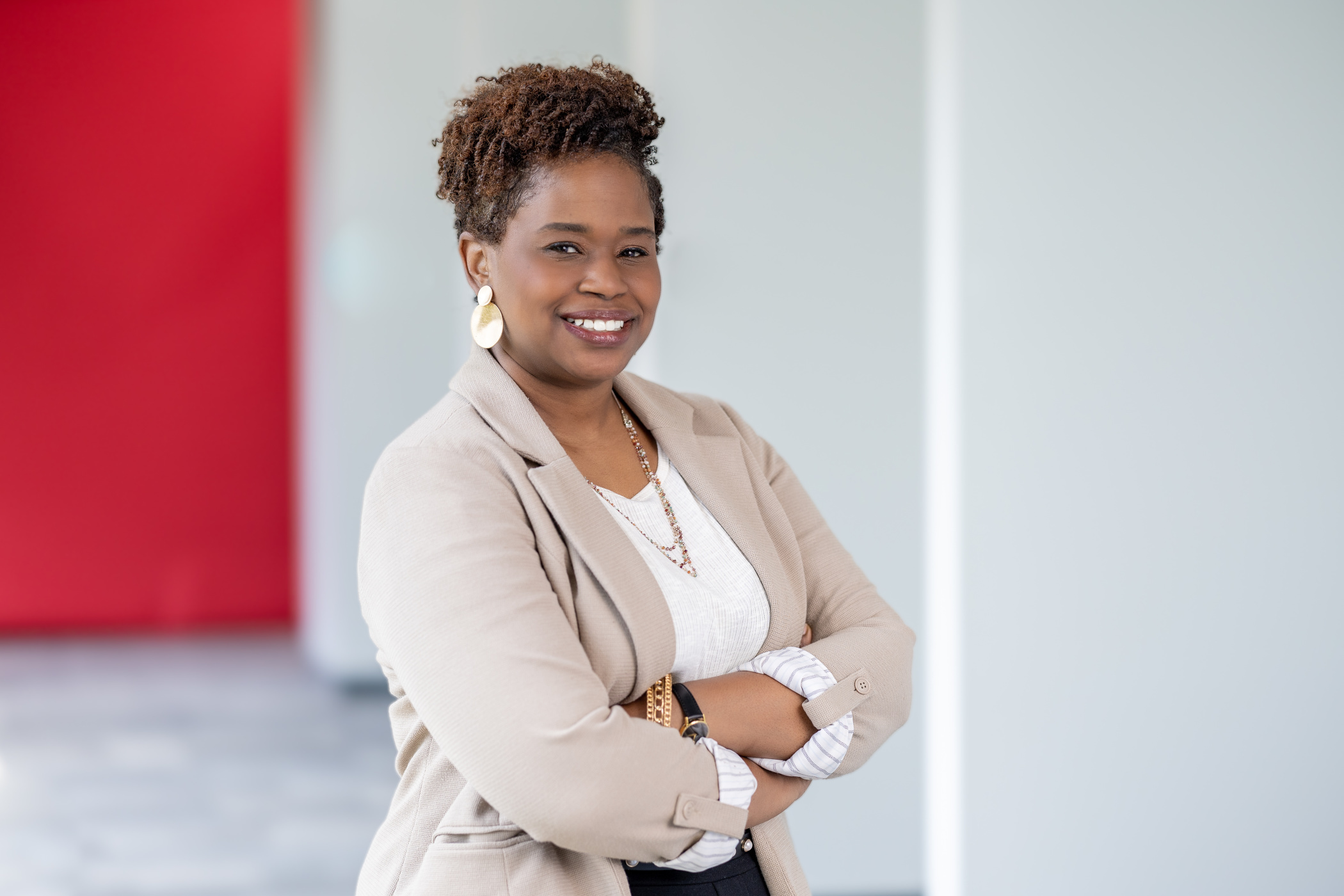 Vanguard Professional Crew Portrait Professional portrait of Rashanda P. standing with arms crossed in a modern office environment. She is wearing a light beige blazer over a white top with layered jewelry and earrings. The background is softly blurred, featuring neutral-colored walls and a red accent panel.
