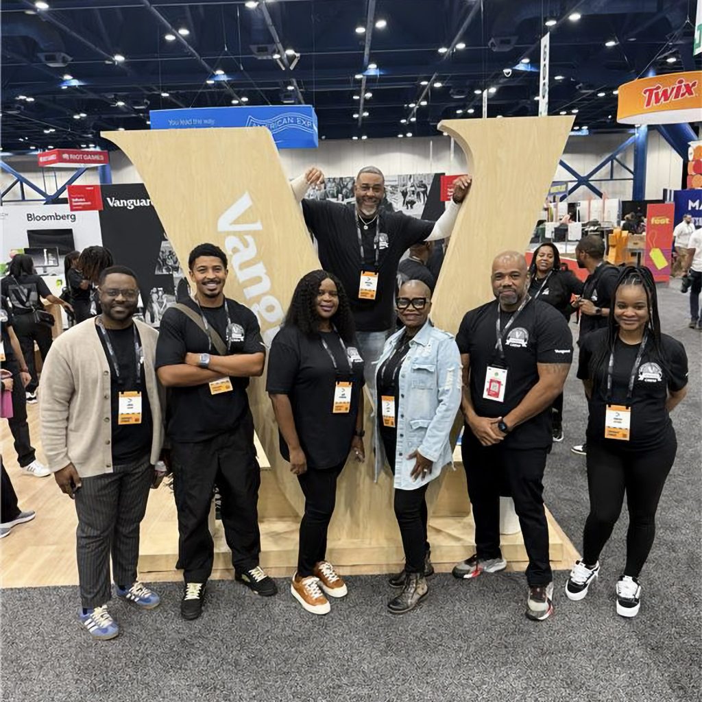 IIolo standing with crew members in the booth in front of large “V” sign at AfroTech conference.
