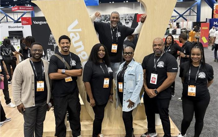 IIolo standing with crew members in the booth in front of large “V” sign at AfroTech conference.