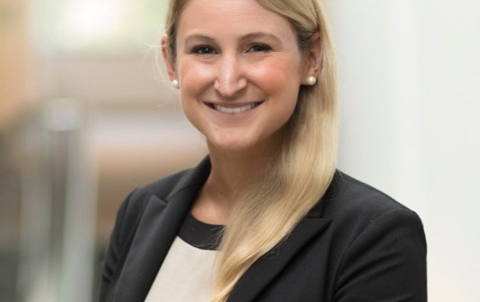 Lauren standing indoors, smiling at the camera in a black blazer and light top, with a blurred hallway in the background.