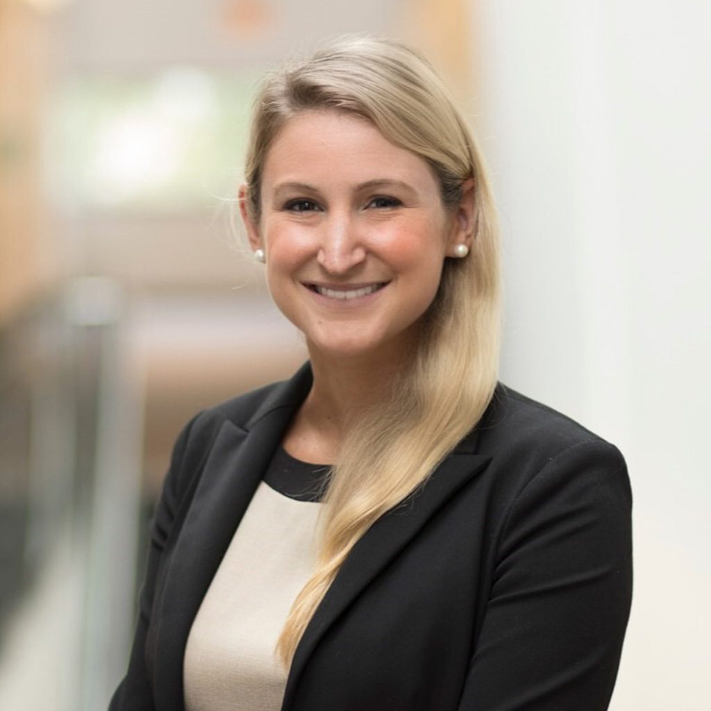 Lauren standing indoors, smiling at the camera in a black blazer and light top, with a blurred hallway in the background.