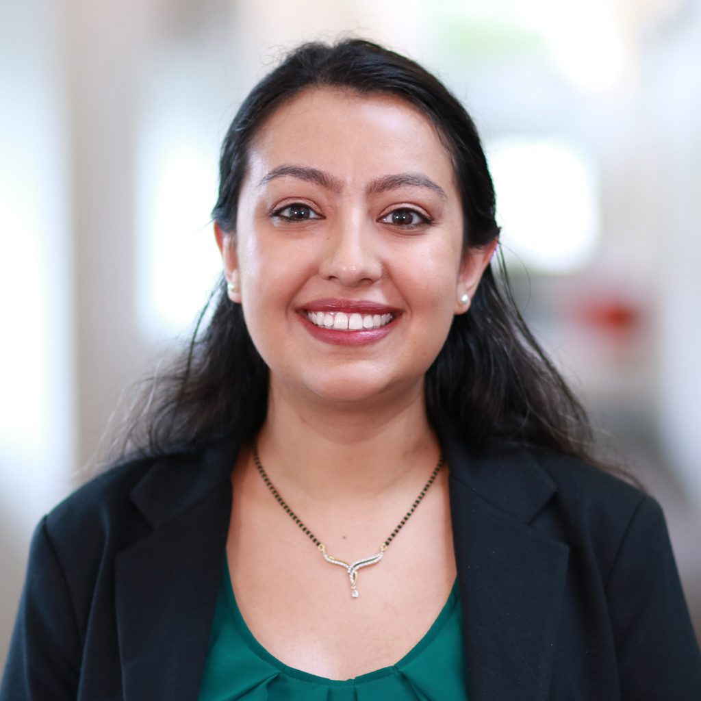 Perryne D. smiling confidently in a green shirt and black blazer. She is against a blurred background of a hallway in an office setting.