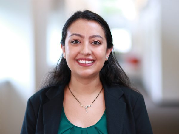 Joy U. smiling in a modern office setting with red and white walls and glass railings, wearing a black blazer over a white top.