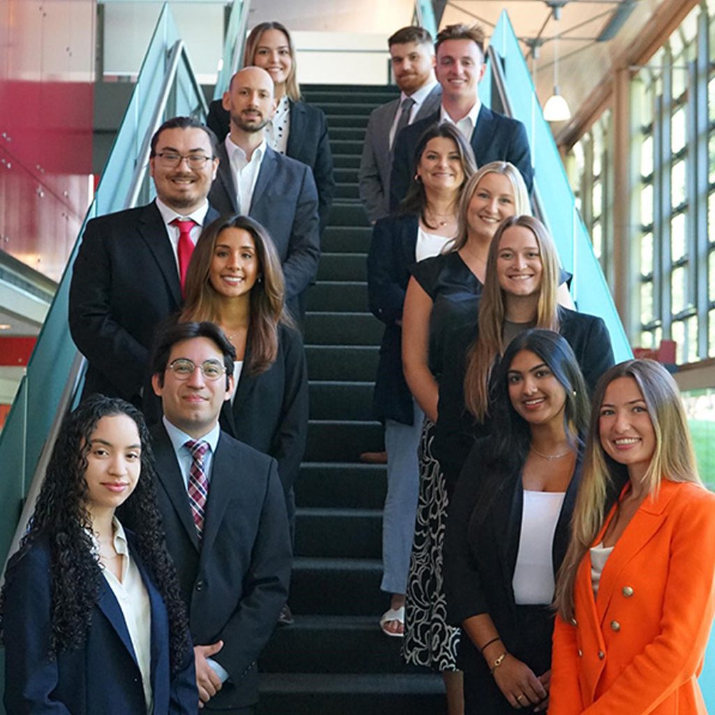 Cohort of crew in a Vanguard specialty program, dressed in business attire and standing on an indoor staircase with glass panels and large windows in the background.