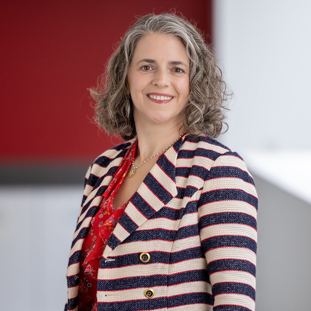 Professional headshot of Liz M. wearing a navy and white striped blazer with gold buttons over a red patterned blouse. The background features a modern indoor setting with a red accent wall and soft natural lighting.