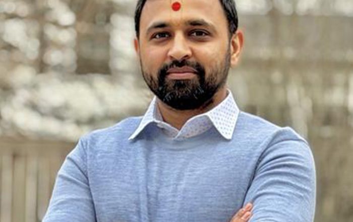 Neel P. standing outdoors with arms crossed, wearing a light blue sweater over a collared shirt, with a wooden fence and trees in the background.