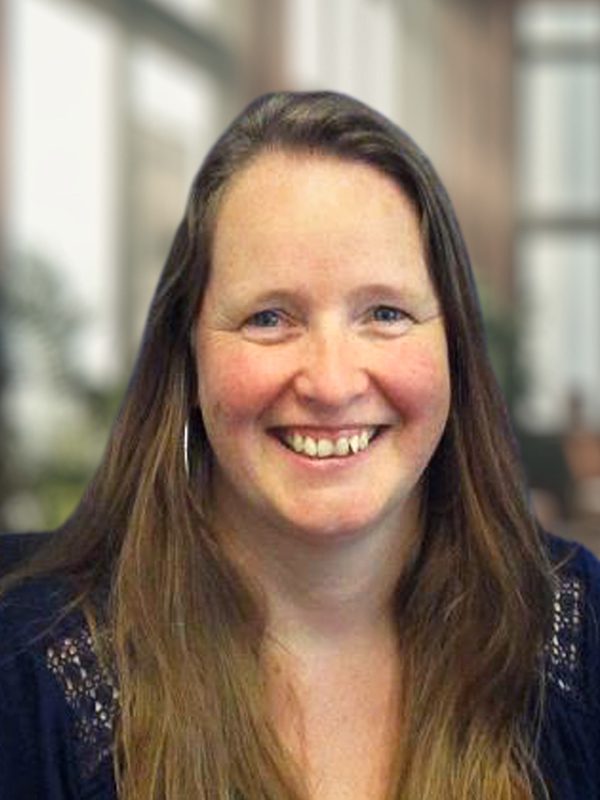 Els smiling in a softly lit setting with large windows and greenery in the background.