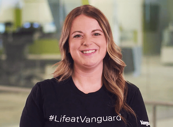Vanguard recruiter standing indoors in a modern office setting, wearing a black T-shirt with the white text “#LifeatVanguard.” The background shows softly focused office furniture and glass walls, with neutral lighting and a professional workplace environment.