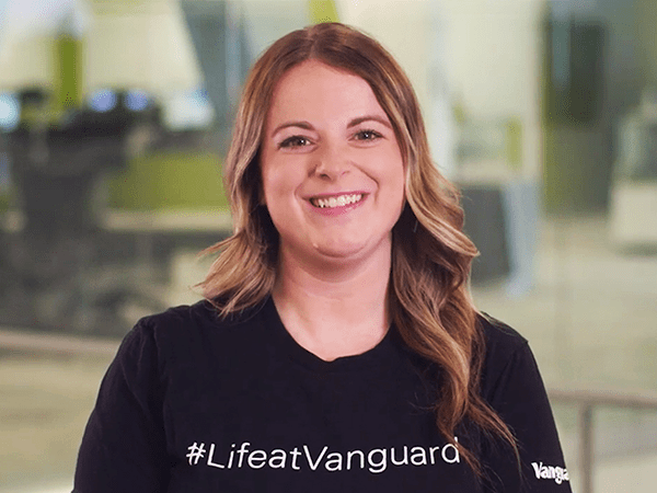 Vanguard recruiter standing indoors in a modern office setting, wearing a black T-shirt with the white text “#LifeatVanguard.” The background shows softly focused office furniture and glass walls, with neutral lighting and a professional workplace environment.