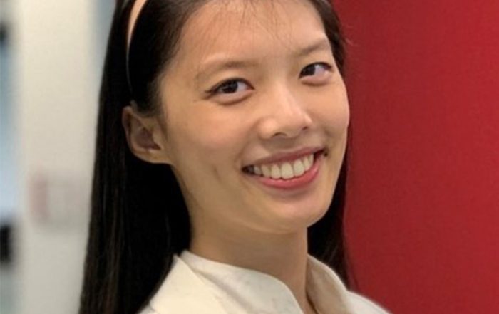 Candy standing indoors in front of a solid red wall, smiling confidently, wearing a light-colored blouse and cardigan.