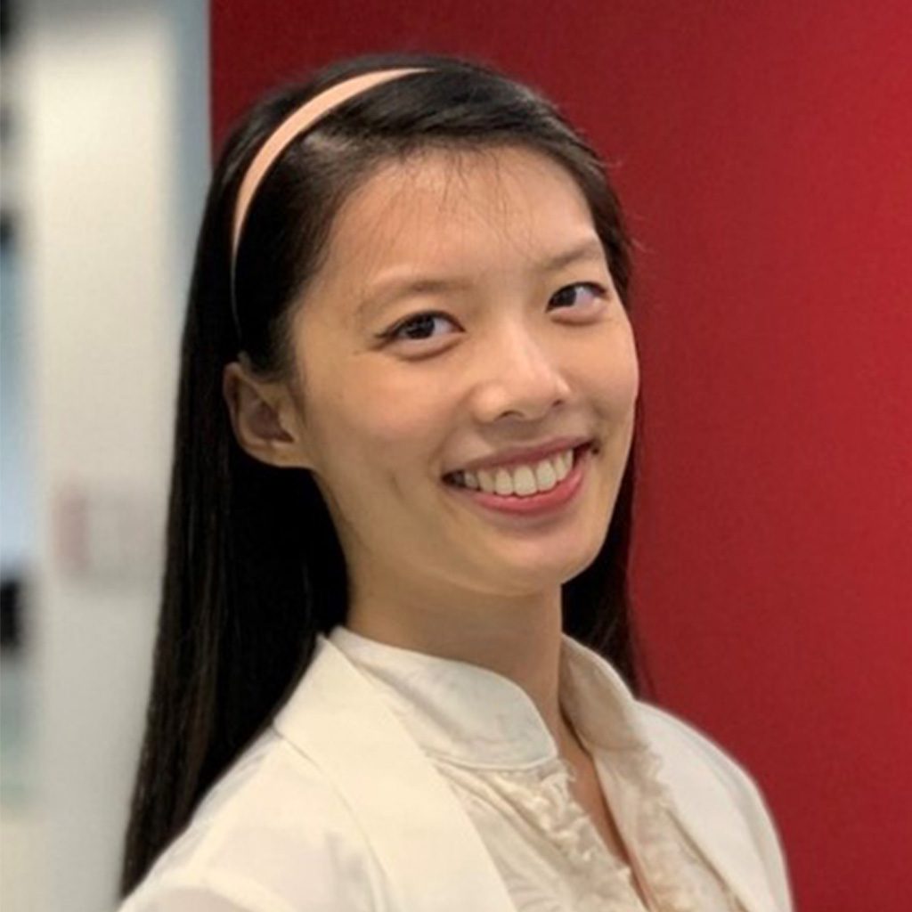 Candy standing indoors in front of a solid red wall, smiling confidently, wearing a light-colored blouse and cardigan.