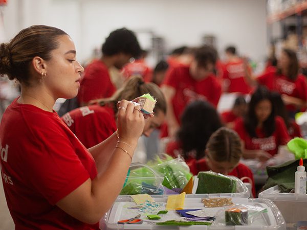 : A group of interns wearing red shirts work at tables in a warehouse workspace, sorting and decorating craft materials inside plastic bins. Shelving, boxes, and supplies fill the background as volunteers work side by side.