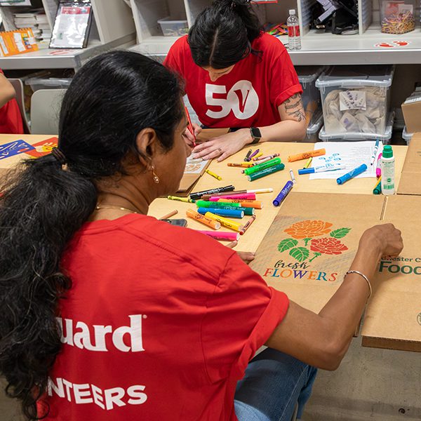 Several interns wearing red “Community 365” shirts sit around a table in a warehouse workspace, drawing and writing on cardboard boxes with markers. Crates of supplies, shelves, and labeled boxes are visible in the background.