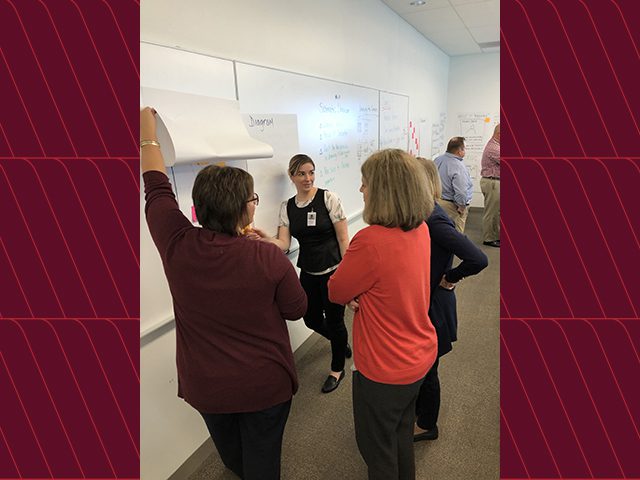 four employees collaborating at work in front of a white board. One employee is holding a page up as they discuss the ideas that are listed on the board.