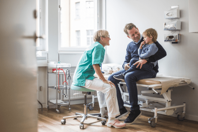 Father holding his young son in a doctor's exam room while a smiling doctor talks to the child.