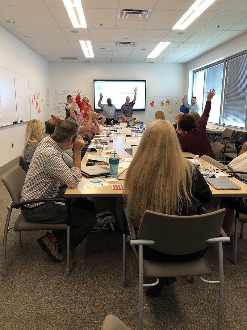 employees during a meeting in conference room. Several employees are raisng their hands in response to a question.