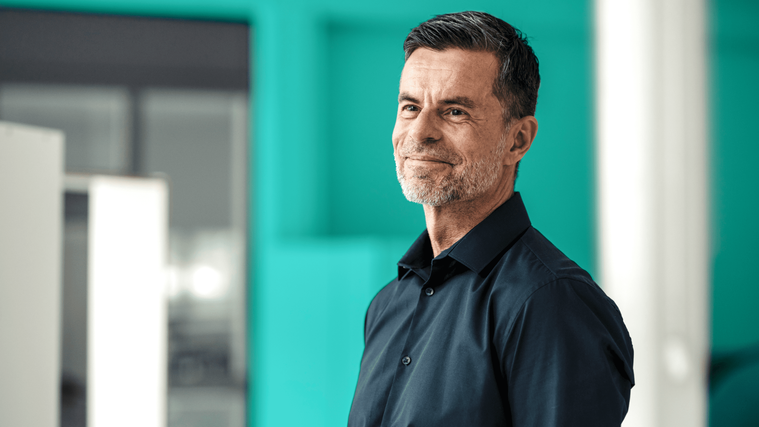 A man looks off into the distance, smiling, in front of a turquoise wall in the office.
