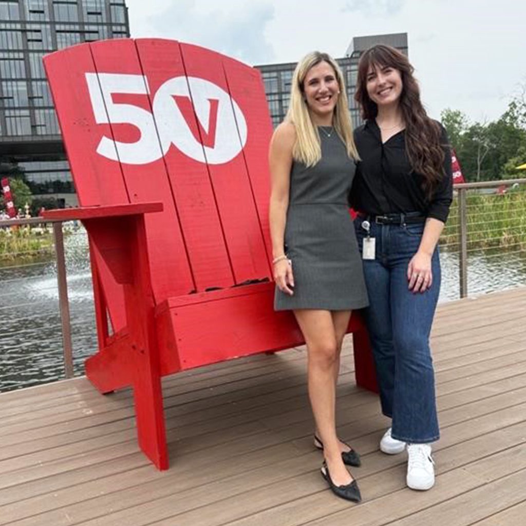 Two women standing on a wooden deck in front of a large red Adirondack chair with '50' painted in white on the backrest, where the '0' contains a stylized letter 'V'. Behind them is a body of water and modern buildings.