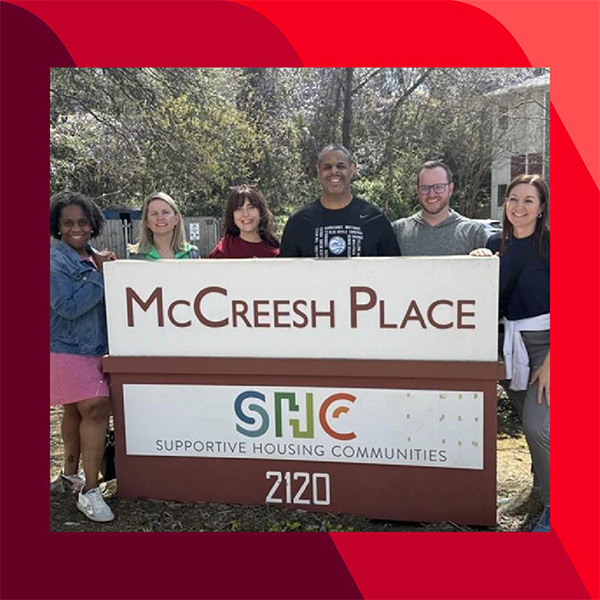 Haley and five other people stand behind a sign that reads "McCreesh Place" and "Supportive Housing Communities," with the address "2120" at the bottom. Trees and buildings are visible in the background