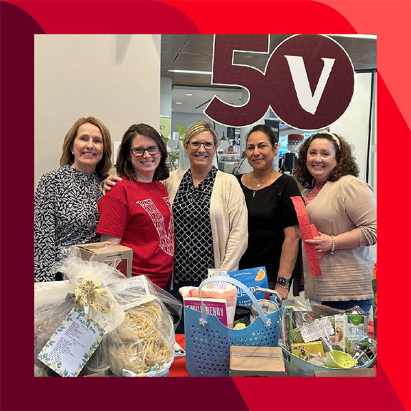 Jill and four other people stand behind a table with various gift baskets, with a large "50V" sign in the background.