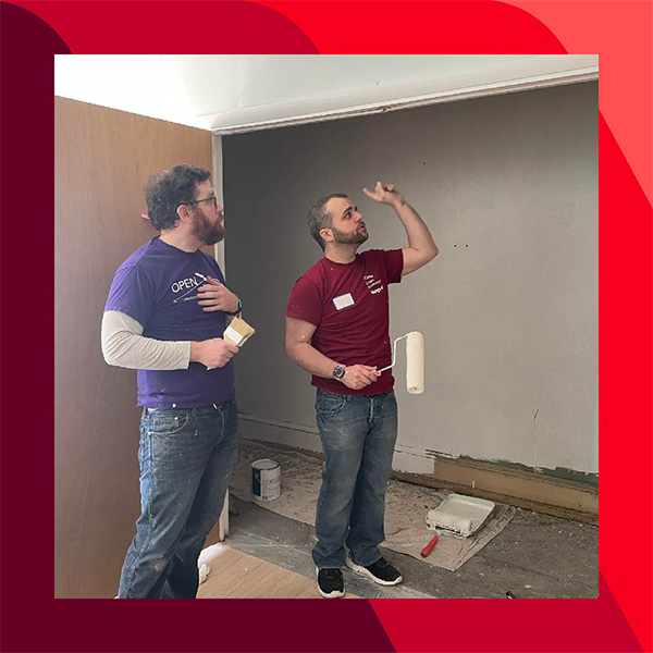 Josh U-A and another person stand in a room being painted, holding a paintbrush and roller, with supplies on the floor and partially painted walls behind them.