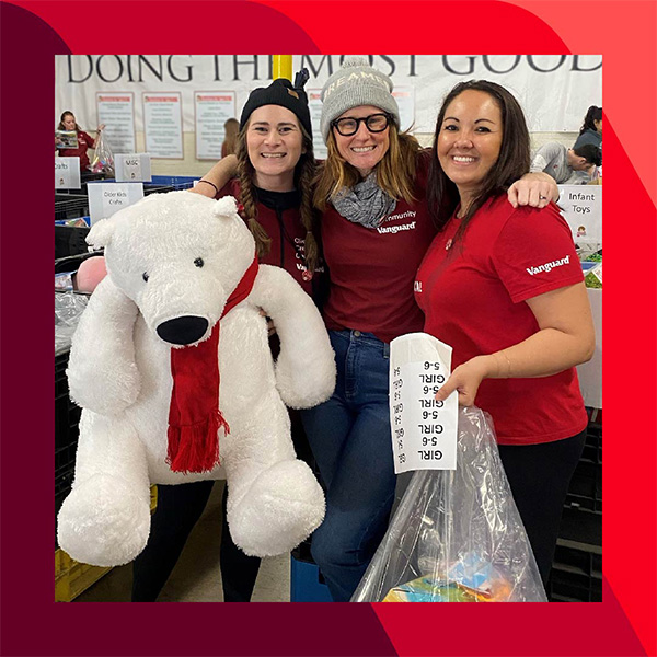 Kim M. in the middle of two other people, arm and arm, wearing red "Vanguard" shirts stand together, holding a white teddy bear and a bag of toys.