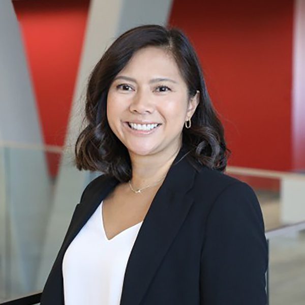 Joy_Umali_Headshot_V2 Joy U. smiling in a modern office setting with red and white walls and glass railings, wearing a black blazer over a white top.