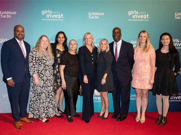A group of nine people standing together on a red carpet in front of a backdrop with the Girls Who Invest and Goldman Sachs logos. Candy Z. is positioned on the far right of the group.