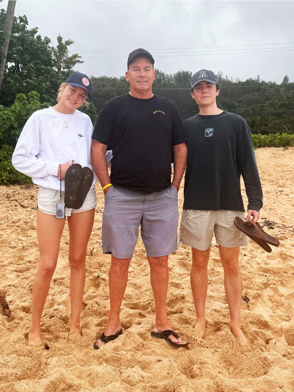 Tracy L.’s daughter, husband, and son standing barefoot on a sandy beach outdoors, wearing casual clothing and baseball caps, with greenery and an overcast sky in the background.