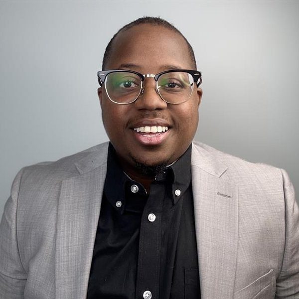 Kenneth in a light gray blazer and black shirt wearing eyeglasses against a neutral background
