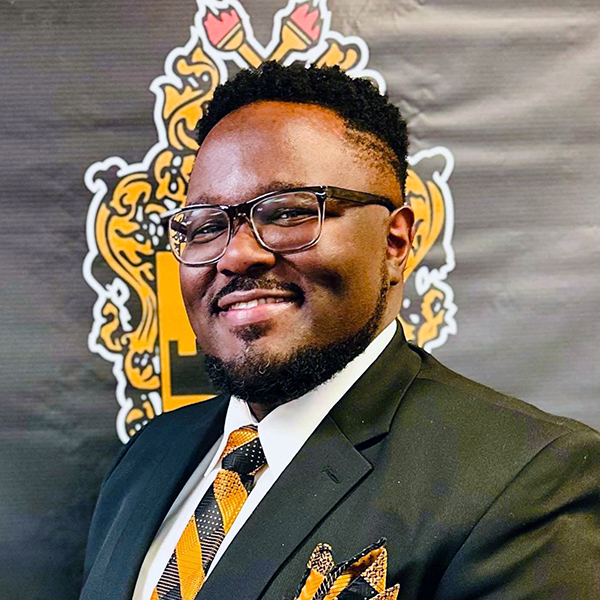 Vanguard_Social_WebContent_CareersBlog_LeadershipDevelopment_Square_ChadL Chad L. wearing a dark suit, patterned tie, and matching pocket square, standing in front of a black-and-gold backdrop.