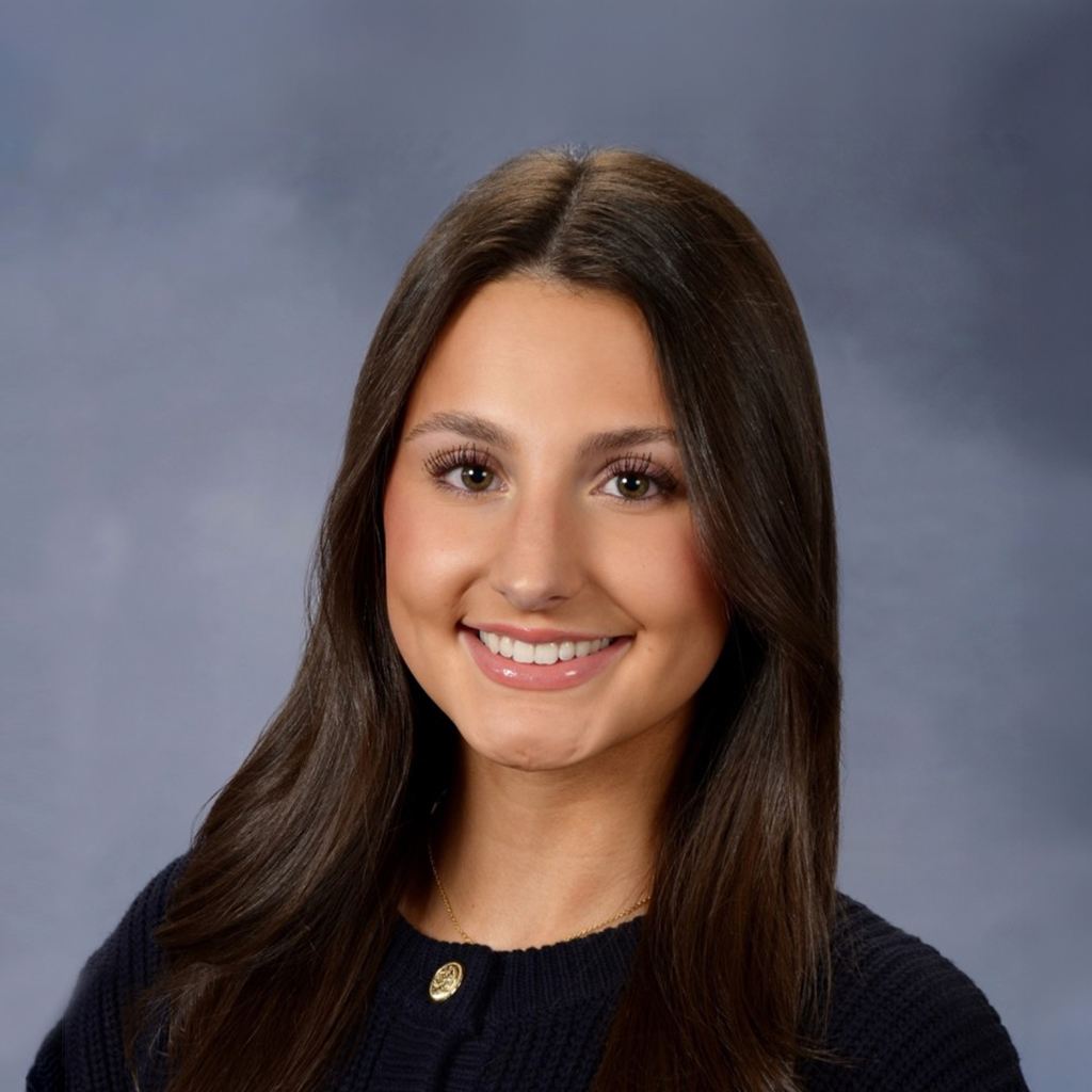 Professional headshot of Rachel W. with long dark hair wearing a dark sweater and a necklace, posed against a gray studio background.